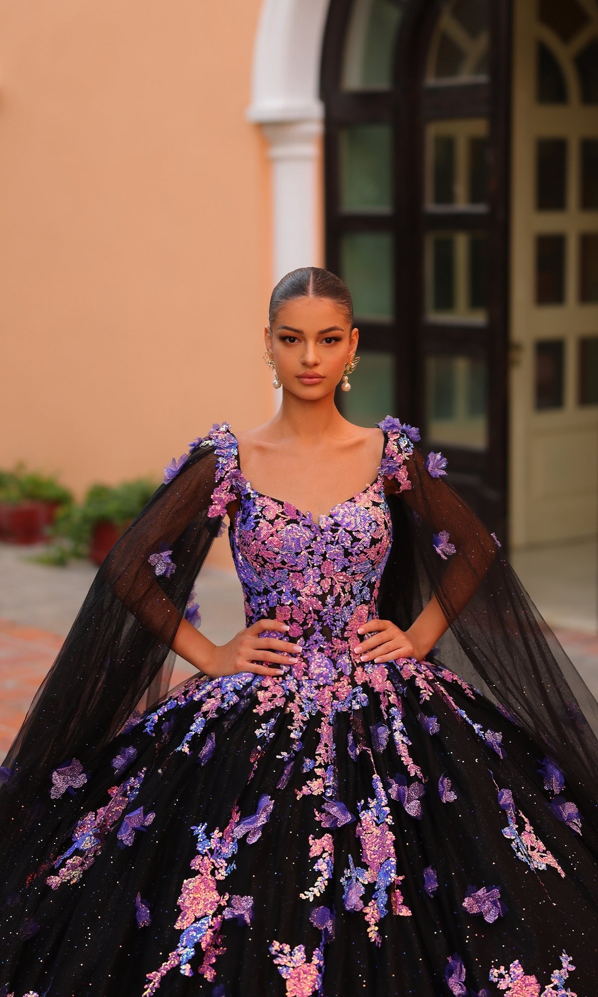 A woman stands outdoors in front of a building, wearing the Quinceanera Dress 54318 by Amarra?a black ball gown with pink and purple floral embroidery and sheer black sleeves?her hands resting on her waist.